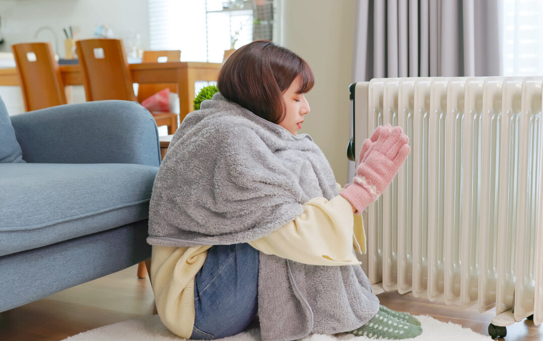 Woman wrapped in a blanket next to a radiator in a living room, trying to stay warm.